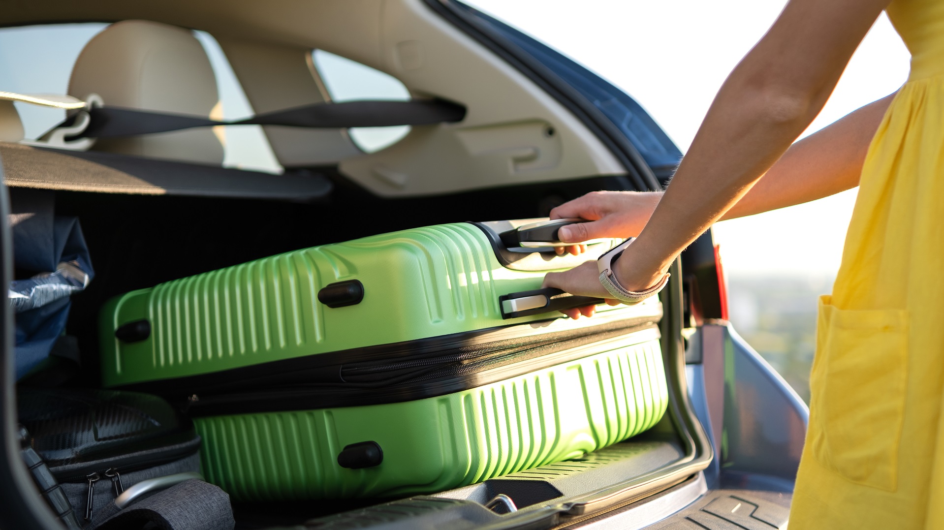 Young woman in yellow summer dress taking green suitcase from car trunk. Travel and vacations concept.